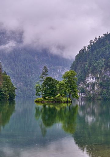 Serene view of a small island with trees reflected in a calm lake, surrounded by misty mountains and cloudy sky in a tranquil landscape.