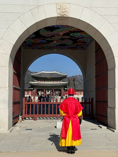 Traditional guard in colorful attire stands at Gyeongbokgung Palace gate in Seoul, South Korea, with vibrant architecture in background.