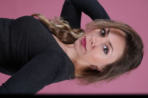 A beautiful Caucasian woman with blonde hair wearing a black blouse posing against a pink background