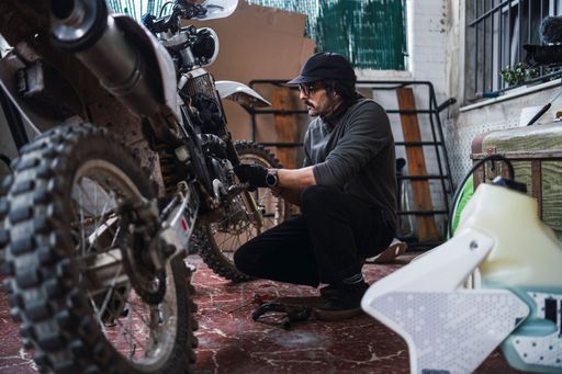 Middle-aged man repairing a motorcycle in a garage, wearing casual attire and a cap, showcasing a hands-on approach to maintenance.