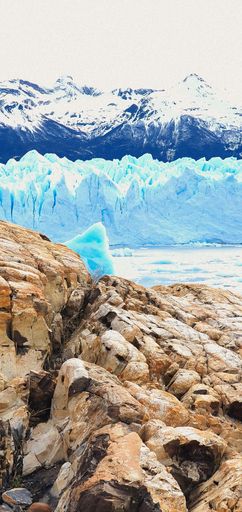 A vertical shot of rocks on the shore of a lake with glaciers and snowy mountains