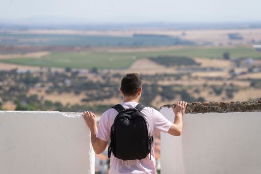 Young Caucasian man in shorts and pink short-sleeved T-shirt with backpack with his back to the horizon leaning against a wall.