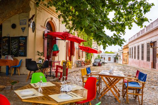 Charming outdoor cafe with colorful chairs and tables on a cobblestone street in Colonia del Sacramento, Uruguay, under lush green trees.