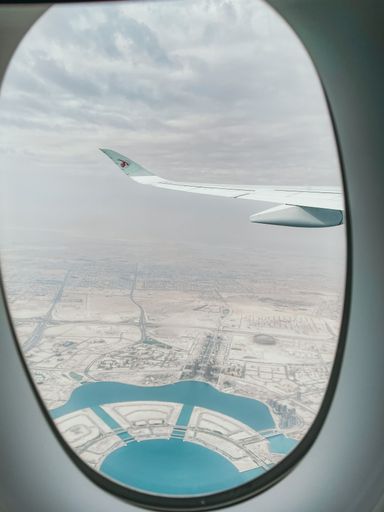 View from an airplane window showing a cityscape with a wing and a coastal area below under a cloudy sky.