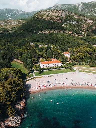 Aerial view of a scenic beach with turquoise waters and lush greenery surrounding a historic building, people enjoying a sunny day.