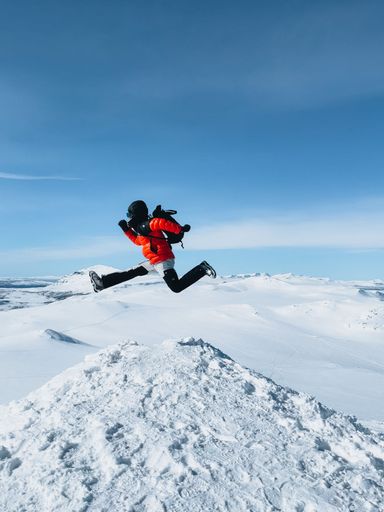 A person in a red jacket jumps energetically in a snowy mountain landscape under a clear blue sky.