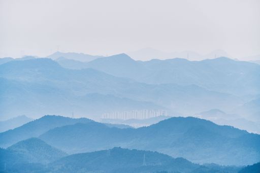 A serene view of misty blue mountains with a distant city skyline under a soft sky, showcasing natural beauty and urban contrast.