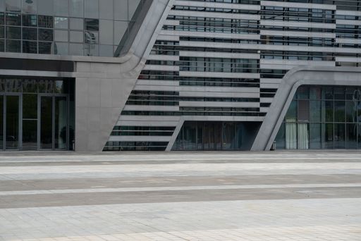 Modern architectural facade with geometric lines and glass panels, featuring a spacious plaza in the foreground.
