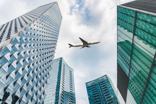 An airplane flying over modern skyscrapers in a cityscape view with blue sky and clouds.