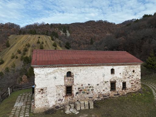 A weathered stone church with a red roof stands against rolling hills, exuding history and serenity.