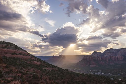Scenic view of Sedona's red rock formations under a dramatic cloudy sky with sun rays breaking through, capturing the natural beauty of Arizona.