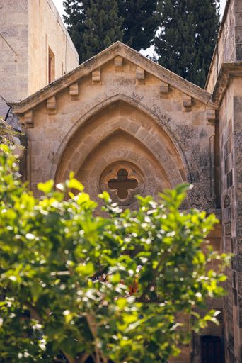 Historic stone building facade with gothic arch and lush greenery in foreground, under a sunny sky.