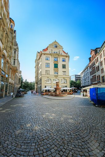 A scenic view of a European city square with a historic building and fountain under a clear blue sky.