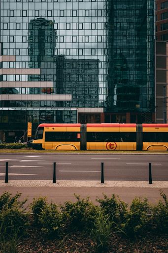 A modern yellow tram passing by an urban cityscape with glass buildings reflecting the skyline on a cloudy day.