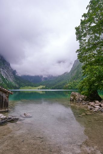 Scenic view of a serene lake surrounded by lush green forests and misty mountains under a cloudy sky.