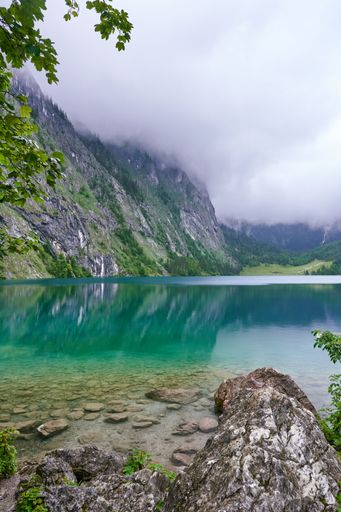 A serene view of an alpine lake with clear turquoise water and misty mountains in the background.