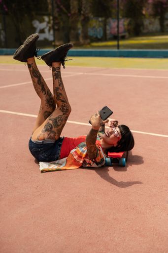 A vertical shot of a Spanish Caucasian girl with tattoos and short black hair, lying on a skateboard and taking a selfie outdoors