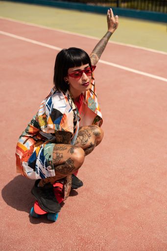 A vertical shot of a Spanish Caucasian girl with tattoos and short black hair, posing with a skateboard outdoors