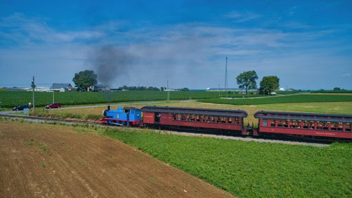 A Thomas the tank engine pulling passenger cars and blowing smoke on a beautiful sunny day in Ronks