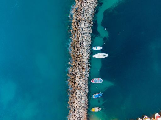 Aerial view of boats anchored along a rocky breakwater in clear blue waters, showcasing a serene coastal scene.