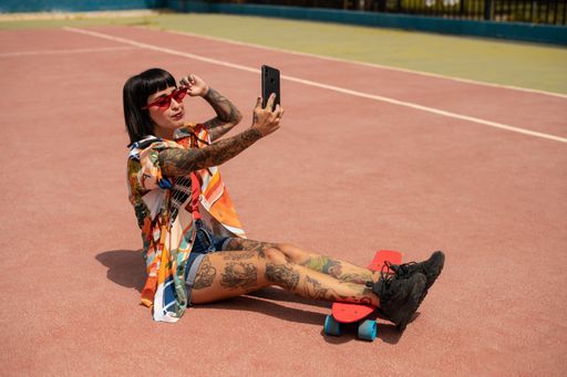 A shallow focus shot of a Spanish Caucasian girl with tattoos and short black hair, lying on a skateboard and taking a selfie outdoors