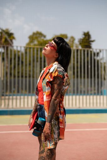 A vertical shot of a Spanish Caucasian girl with tattoos and short black hair, holding a skateboard outdoors