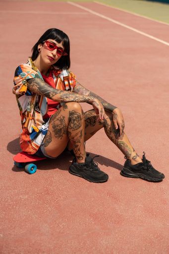 A vertical shot of a Spanish Caucasian girl with tattoos and short black hair, sitting on a skateboard outdoors