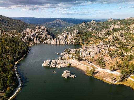 Scenic aerial view of Sylvan Lake surrounded by rocky formations and lush forests in Custer State Park, South Dakota, under a partly cloudy sky.