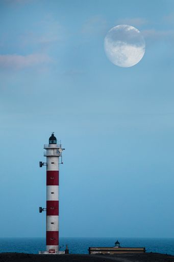 A vertical shot of a lighthouse on the sky background with the moon