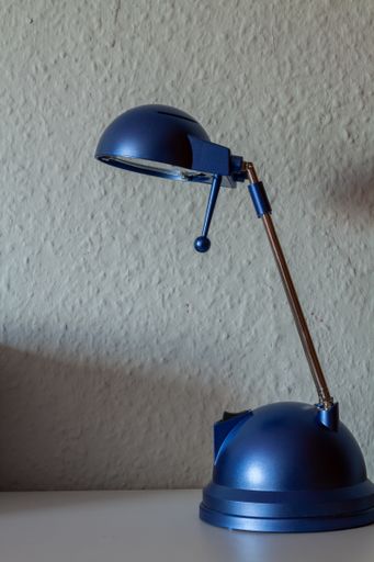 A blue metallic table lamp on a table against a white textured wall