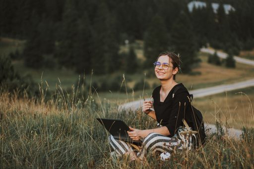 A shallow focus shot of a Bosnian Caucasian woman with a laptop in her lap outdoors in a field, looking happy