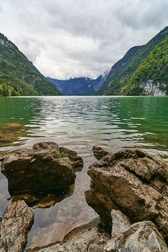Scenic view of a serene lake surrounded by lush green mountains under a cloudy sky, with rocks in the foreground.