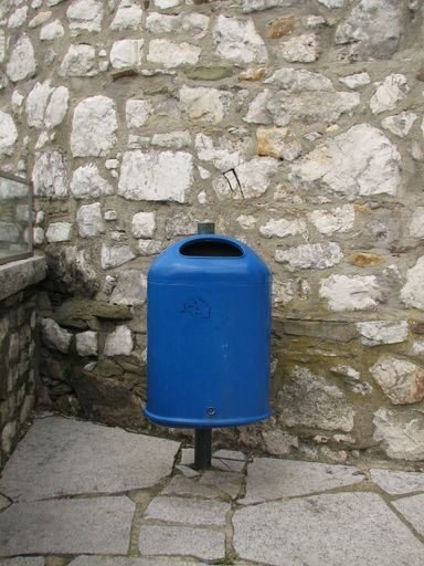 A view of a blue trashcan on a pole in the street near the stone wall in the park