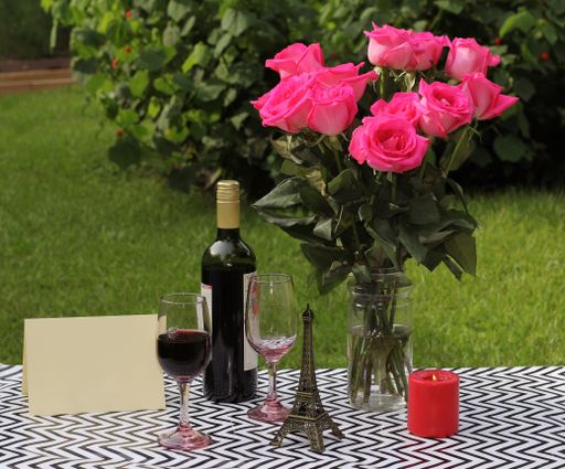 A view of a bouquet of roses in a glass vase with a bottle of wine, two glasses, a candle, statuette, and postcard on the ground