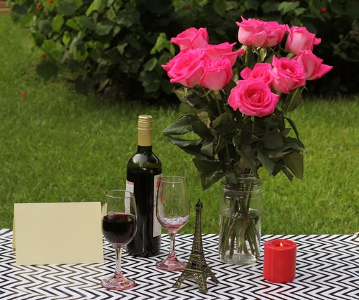 A view of a bouquet of roses in a glass vase with a bottle of wine, two glasses, candle, statuette, and postcard on the ground