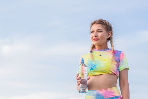 Young beautiful girl in bright tracksuit stands against background of blue summer sky. He holds plastic bottle with drinking water in his hand.
