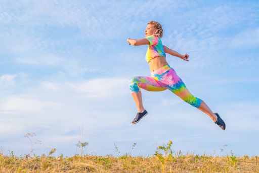 Young woman in a bright light tracksuit flies in a long stride over the meadow against the background of the blue summer sky. Outdoor sports.