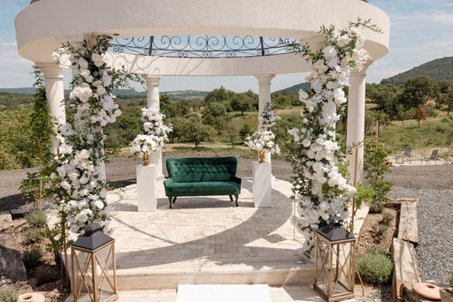 Elegant outdoor wedding gazebo adorned with white flowers and a green sofa, set in a scenic landscape under a clear blue sky.