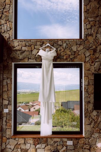 Elegant white wedding dress hanging in front of a large window with a scenic view of a countryside landscape and blue sky.