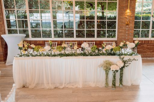 Elegant wedding reception table with floral arrangements and white linens in a bright, airy venue with brick walls and large windows.