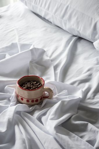 A cozy scene with a coffee cup filled with coffee beans on a rumpled white bedsheet, creating a warm and inviting atmosphere.