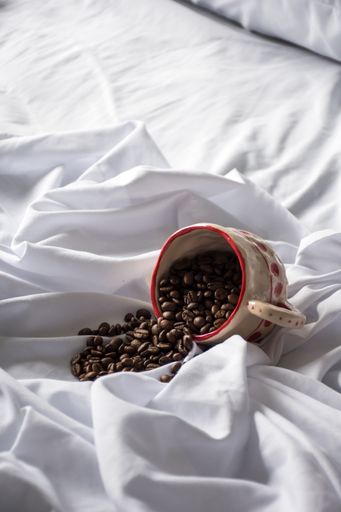A cozy scene with a coffee mug filled with coffee beans spilling out of its cup on a crumpled white bed sheet.