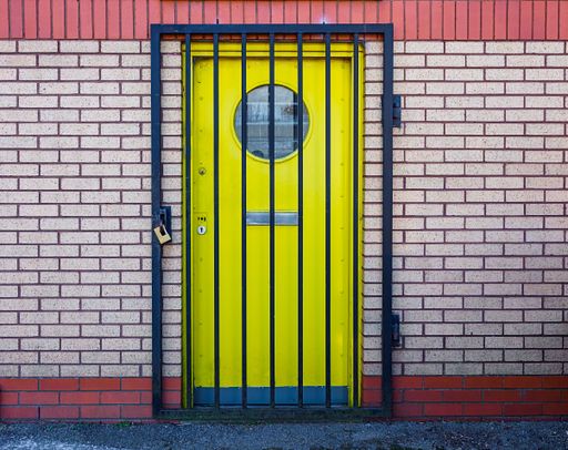 A yellow door with a round window is secured behind black metal bars. The door is set in a brick wall with red and beige bricks.