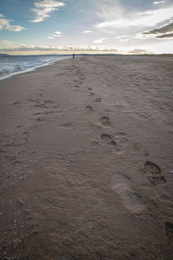 Footprints on a sandy beach at sunset with a distant figure walking along the shore under a partly cloudy sky.