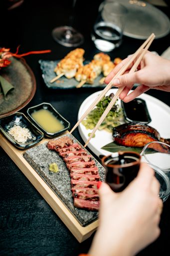 Close-up of a person using chopsticks to pick up sliced beef from a stone platter, surrounded by various Japanese dishes on a dark table.