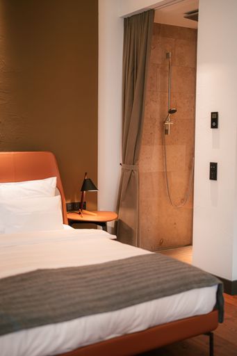 Modern hotel room interior with a cozy bed, bedside table, and a view into a stylish bathroom with a shower.