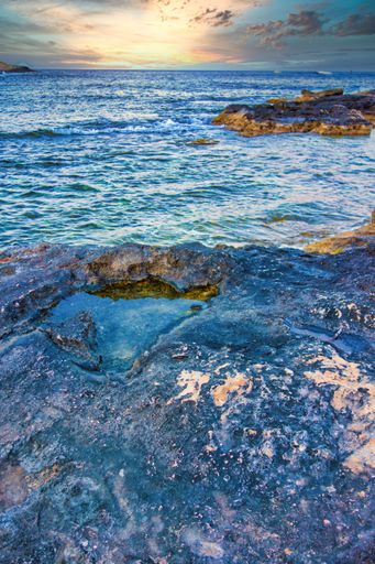 A vertical shot of the waves hitting the rocks on the coast during the sunset.