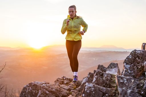 A woman in her 30s jogging on rocky terrain during sunset with mountains in the background, wearing a yellow jacket.