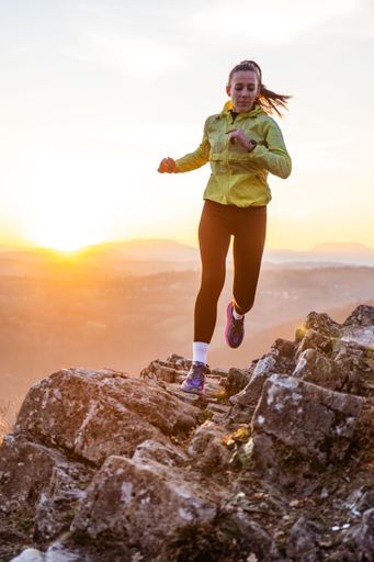A woman in her 20s running on rocky terrain at sunset, wearing athletic gear