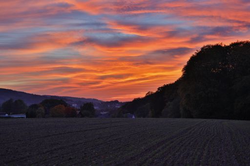 A landscape of a field surrounded by greenery and hills under a cloudy sky during a vibrant sunset
